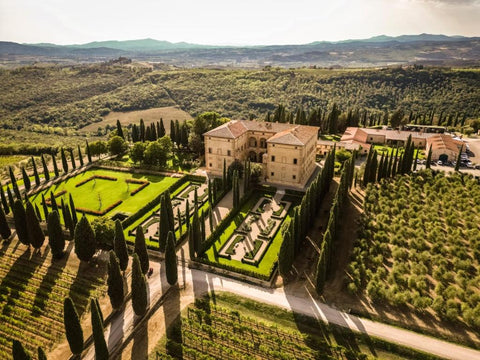 Aerial view of the Argiano Winery Estate with formal gardens and tall cypress trees, surrounded by vineyards and olive groves in a rolling Tuscan landscape.