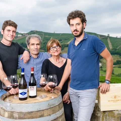 A group photo of The Bera Family standing in front of a barrel with wine glasses and bottles on the top. Scenery of their windery fields can be seen in the background. 