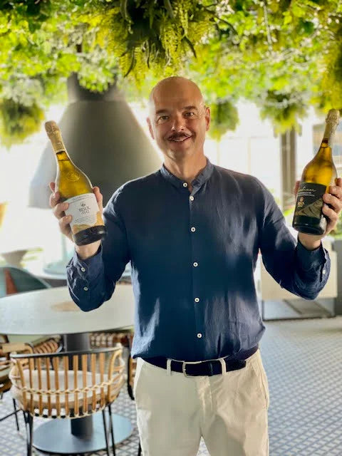 A photograph of Gianluca Bisol wearing a navy shirt while holding two wine bottles from the Bisol Winery. The scene is lit with soft and bright lighting while greenery surrounds the top.