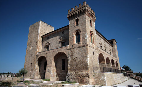 A large historic stone building located in the Feudi Bizantini Winery. It features arched ground-floor openings and a crenellated tower, photographed outdoors against a clear blue sky.