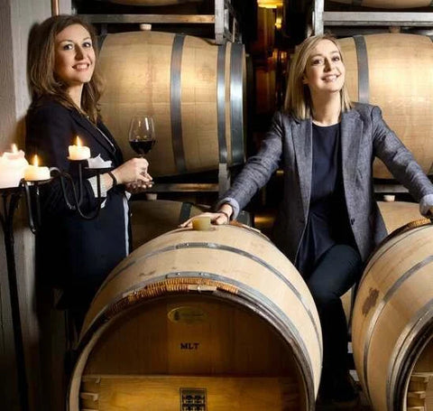 Two women in formal attire are positioned between large wine barrels in the ll Barroccio Winery building. More rows of wine barrels are seen behind them.