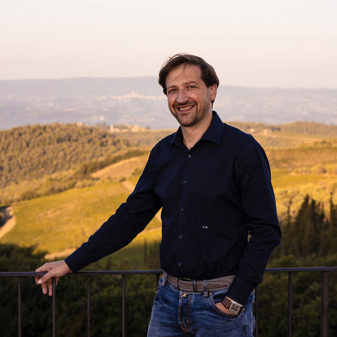 A scenic portrait of Emanuele Reolon standing in front of a metal fence wearing a navy long sleeve shirt. Behind him is land a part of the Isole e Olena Winery. 